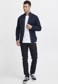 Jersey de punto azul marino con cremallera y tejido texturizado, llevado sobre una camiseta blanca, combinado con jeans negros y zapatillas en negro y blanco.