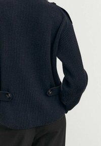 Pull en maille côtelée bleu marine à manches longues, avec des accents de boutons à la taille et à l'épaule. Texture douce et coupe décontractée.
