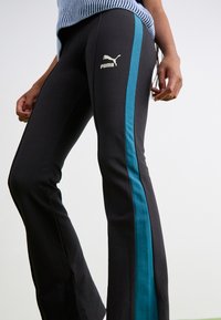 Čierne športové nohavice s tyrkysovými bočnými pruhmi, vyrobené z mäkkej tkaniny. Obsahujú malé zlaté logo Puma na stehne. Dizajn so rozšírenými nohavicami.
