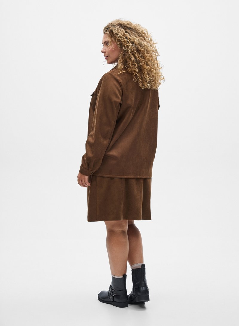Frau mit lockigem Haar, die eine braune Wildlederjacke und einen Rock trägt, kombiniert mit schwarzen Stiefeletten, vor einem schlichten weißen Hintergrund steht.