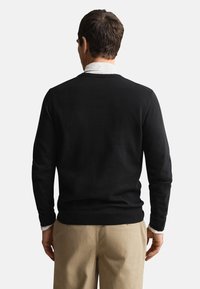 Schwarzer Strickpullover mit Rundhalsausschnitt, langen Ärmeln sowie gerippten Bündchen und Saum. Über einem hellfarbenen Hemd mit Kragen getragen, zeigt eine glatte Textur.