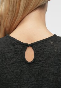 Czarna bluzka o teksturowanej powierzchni z okrągłym dekoltem, posiadająca wycięcie w kształcie dziurki na plecach, zakończona małą pętlą z tkaniny do zapięcia.