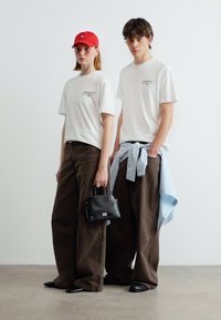 Δύο άτομα με λευκά μπλουζάκια και φαρδιά καφέ παντελόνια· το ένα φορά κόκκινο καπέλο και κρατάει μαύρη τσάντα, ενώ το άλλο έχει μπλε πουκάμισο δεμένο γύρω από τη μέση.