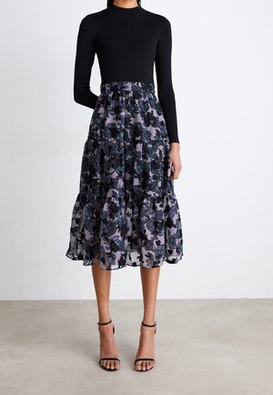 Mujer con top de manga larga negro y falda de longitud media con patrones florales oscuros, combinada con tacones negros con pulsera en el tobillo.