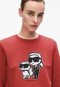Felpa rossa con una grafica di un cartone animato che raffigura una figura con occhiali da sole accanto a un gatto con occhiali da sole, entrambi contornati in nero.