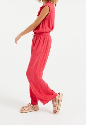 Kvinde iført en lys koralfarvet ærmeløs jumpsuit og lyserøde sandaler med spænder, der går mod en ensartet hvid baggrund.