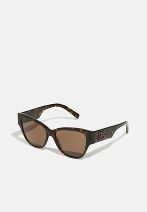 Brązowe okulary przeciwsłoneczne w tortoiseshell o kwadratowym kształcie, z brązowymi szkłami i akcentem logo na zauszniku. Charakteryzują się grubymi oprawkami i gładką teksturą.