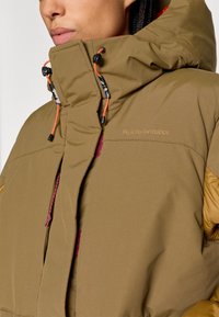 Isoleret hættetrøje i olivengrøn med orange snore. Den har en blød tekstur og kontrasterende gule accenter, mærket med "Peak Performance."