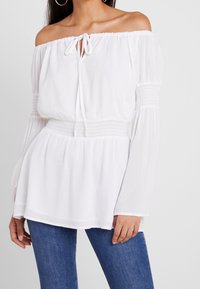 Blouse blanche à épaules dénudées avec un détail à nouer, taille smockée, manches longues et tissu doux. Associée à un jean skinny bleu.