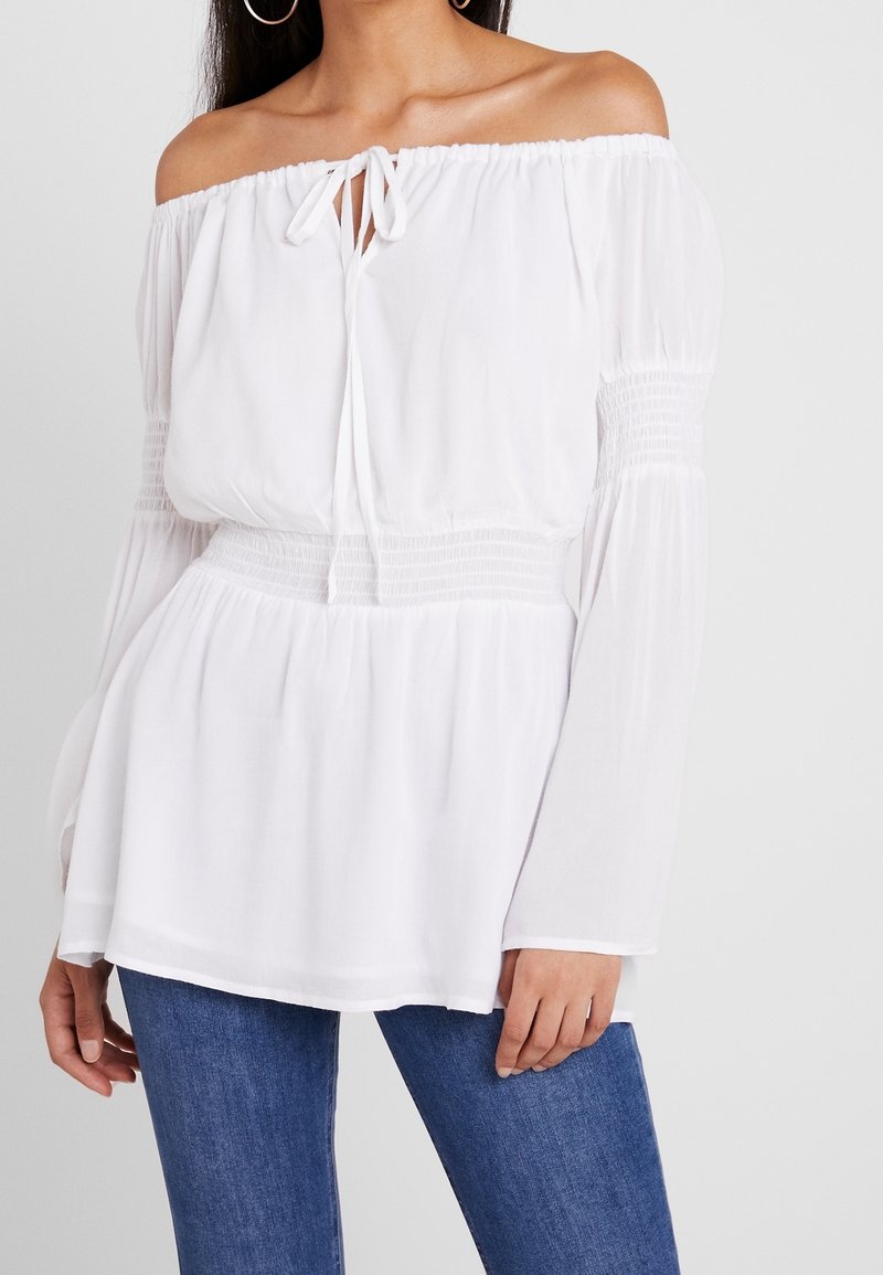 Blouse blanche à épaules dénudées avec un détail à nouer, taille smockée, manches longues et tissu doux. Associée à un jean skinny bleu.