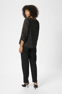 Chemisier noir transparent à pois blancs, manches trois-quarts et coupe décontractée, associé à un pantalon noir ajusté et des chaussures à talons noires.