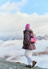 Puchowa brązowa kurtka, białe spodnie i brązowe buty trekkingowe. Różowa czapka i kolorowy plecak z niebieskimi, różowymi i żółtymi akcentami. Górskie tło.