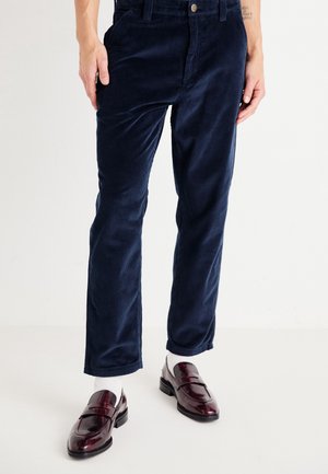 Persoon die marineblauwe corduroybroek, witte sokken en glanzende donkerrode leren loafers draagt, staand tegen een effen lichte achtergrond.