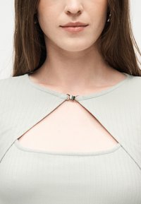 Κοντινό πλάνο μιας γυναίκας που φοράει ανοιχτό γκρι ριγωτό τοπ με ένα μικρό ασημένιο κούμπωμα στο στήθος, που παρουσιάζει ένα τριγωνικό άνοιγμα κάτω από το κούμπωμα.