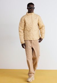 Beige vadderad jacka med hög krage och blixtlås, bärs över beige byxor. Modellen har solglasögon och står mot en neutral bakgrund.