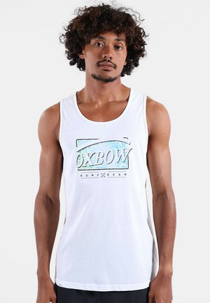 Homme aux cheveux bouclés portant un débardeur blanc avec le texte « OXBOW SURF GEAR » et un graphisme bleu à l'avant, debout devant un fond uni.