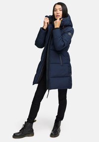 Marineblå puffjakke med høj krave, frontlyfter og justerbar hætte. Har polstrede sektioner og lynlåslommer i siden. Sorte leggings og støvler.