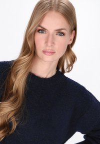 Mørkeblå strikket sweater med rund hals og blød tekstur, båret af en model med langt, bølget hår foran en ensfarvet baggrund.