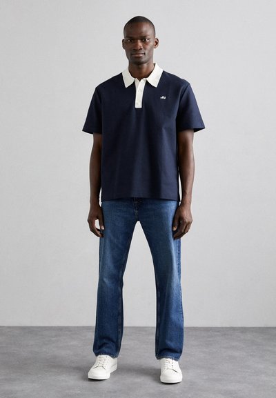 Marineblå polo med hvit krage, korte ermer og en liten logo på brystet, kombinert med lyseblå jeans og hvite joggesko.