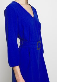 Blå klänning med v-ringning och tre fjärdedelsärmar, utrustad med ett bälte med fyrkantig spänne och texturerad tyg.