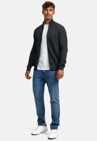 Chaqueta gris con un patrón en zigzag y cremallera sobre una camiseta blanca, combinada con jeans azules y zapatillas blancas.