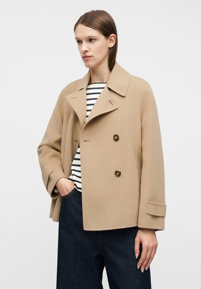 Γυναίκα που φοράει ένα μπεζ διπλοστέλιδο παλτό πάνω από μια ριγέ ασπρόμαυρη μπλούζα και σκούρο μπλε τζιν, ποζάροντας με το ένα χέρι στην τσέπη.