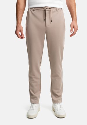 Man met beige, gestructureerde joggerbroek met aantrekkoord in de taille en witte sneakers, staand tegen een effen witte achtergrond.