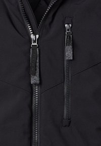 Sort stofjakke med to lodrette metalzips, der har sorte og grå trækstænger, en større lynlås i midten og en mindre lynlås på brystet.