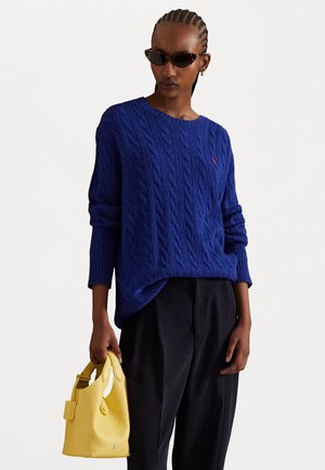 Maglione blu a trecce con maniche lunghe, abbinato a pantaloni neri e una borsa gialla con texture. Gli occhiali da sole completano il look.