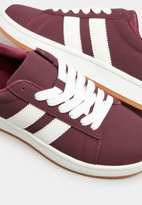 Sneaker bordeaux con design a tre strisce bianche, punta tonda, lacci bianchi e suola in gomma naturale; materiale liscio con dettagli cuciti.