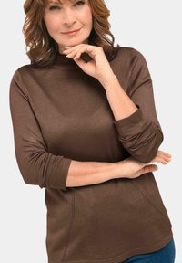 Brauner, langärmeliger Oberteil mit einem mock neckline, das über eine gerippte Textur, seitliche Stickdetails und eine lockere Passform verfügt. Kombiniert mit blauen Jeans.