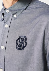 Skjorta med knappar i ljusblå chambray, med vita knappar, en kontrasterande krage och en blå broderad logotyp på vänster sida.