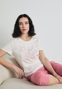 T-shirt in cotone bianco sporco con disegno floreale rosa. Indossato con pantaloni corti rosa chiaro. Modella seduta su una superficie beige di tessuto.