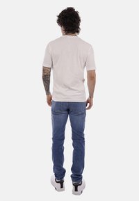 Homme aux cheveux bouclés et tatouages sur le bras droit, portant un t-shirt blanc, un jean bleu et des baskets blanches, debout de dos sur un fond blanc.