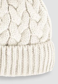 Kapa od vune s pletenim uzorkom u off-white boji, s rebrastim rubom za udoban fit. Mekana, gusta nit pruža toplinu.
