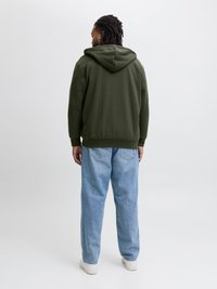 Olivovo zelená mikina s kapucňou, z bavlnenej zmesi, vo voľnom strihu, v kombinácii so svetlomodrými voľnými džínsami a bielymi teniskami.