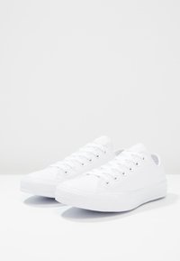Paire de baskets basses blanches propres avec lacets, placées côte à côte sur une surface blanche devant un fond neutre.