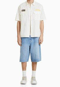 Camicetta bianca a maniche corte con texture e due tasche sul petto; presenta dettagli ricamati neri e gialli. Indossato con pantaloncini di jeans azzurri.