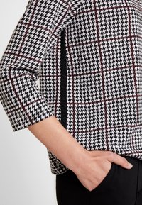 La mano di una donna nella tasca dei pantaloni neri, indossa un top a motivi houndstooth nero, bianco e rosso con maniche a tre quarti e dettagli con spacco laterale.