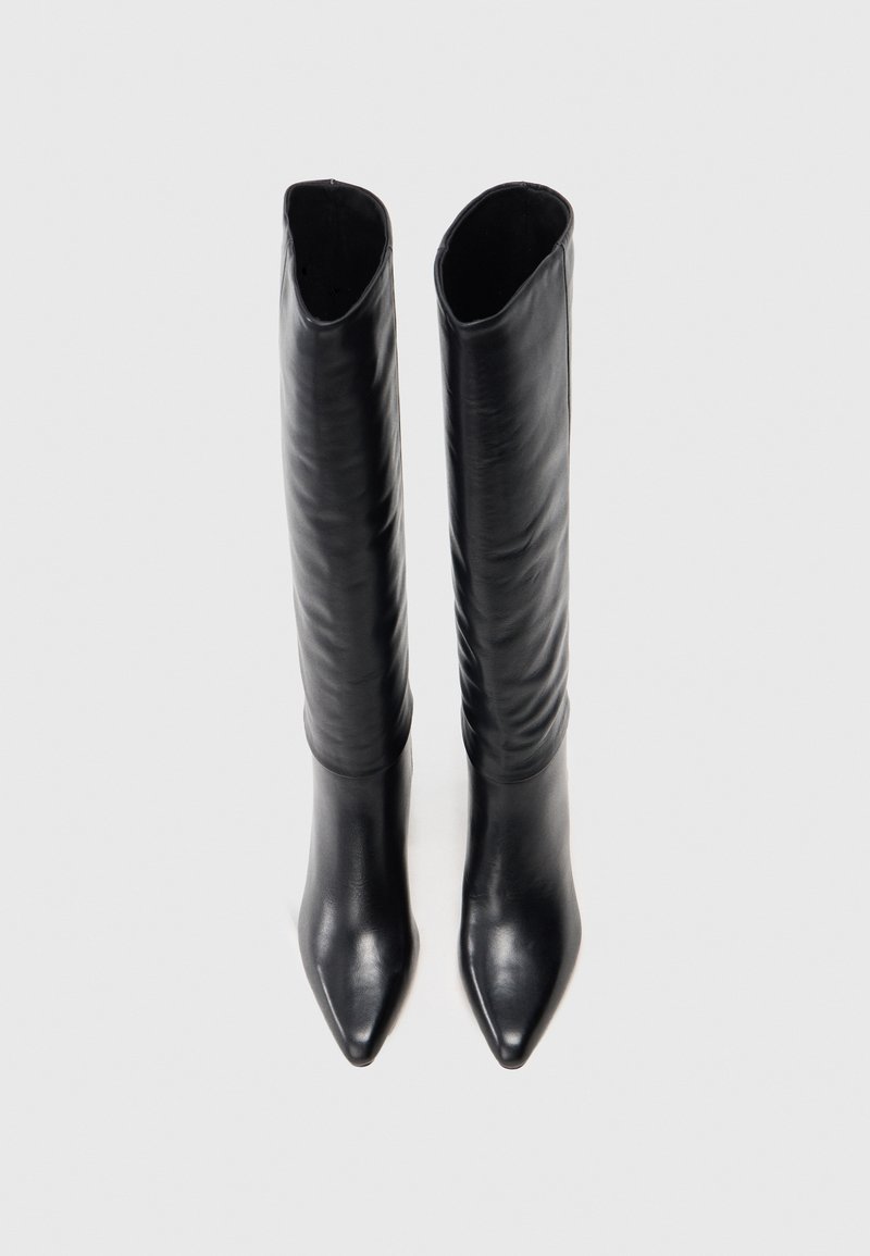 Une paire de bottes noires en cuir hautes avec des pointes accentuées, se tenant droites sur un fond blanc uni.