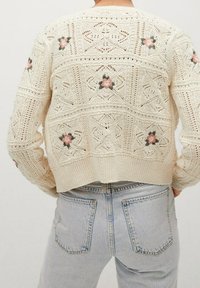 Dzianinowa kremowa kardigan z teksturowym wzorem ażurowym i haftowanymi kwiatowymi detalami w odcieniach różu i zieleni. Krótka długość z ribbowanym wykończeniem.