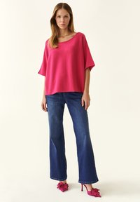 Maglione oversized fucsia in maglia con maniche larghe, abbinato a jeans a campana blu scuro. Le calzature presentano tacchi rosa con dettagli floreali.