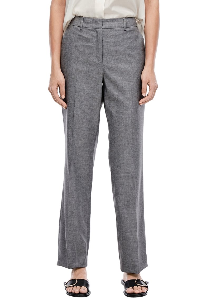 Femme portant un pantalon de tailleur gris droit, une blouse blanche rentrée et des sandales noires avec des boucles dorées, debout devant un fond blanc.