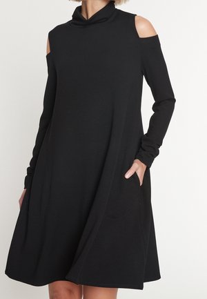 Kobieta ubrana w czarną, długorękawową sukienkę z wycięciami na ramionach, wysokim kołnierzem i bocznymi kieszeniami, stojąca na jednolitym tle.