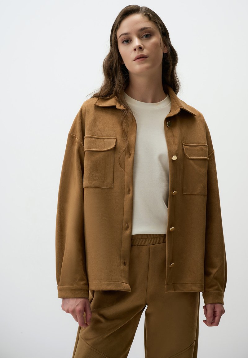 Veste en daim marron avec un devant à boutons, deux poches poitrine et une coupe décontractée, portée sur une chemise crème et un pantalon assorti.