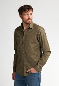 Camisa verde oliva de botones con un sutil patrón geométrico, mangas largas, cuello y corte recto, combinada con jeans azules.