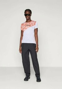 Balta sportinė marškinė su oranžinėmis juostelėmis, juodu logotipu ir juodomis laisvomis kelnėmis, dėvima su akinių nuo saulės ir juodais bateliais.