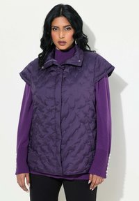 Ärmellwest aus lila Daunenmaterial mit einem gesteppten Muster, Druckknopfkragen und zwei seitlichen Taschen, getragen über einem passenden lila Pullover mit gerippten Ärmeln.