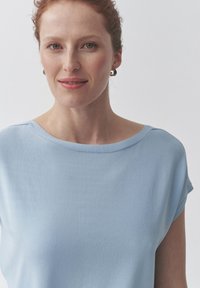 Světle modrý top s krátkým rukávem a širokým lodičkovým výstřihem. Tkanina vypadá jemně a hladce, s jednoduchým, uvolněným designem.