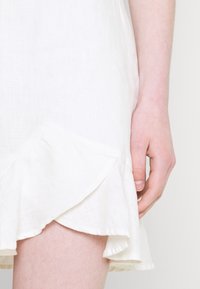Vestido de lino blanco con un dobladillo rizado, mostrado con una mano cerca de él. La textura es ligera y aireada, destacando la suavidad de la tela.